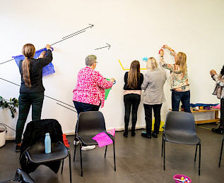 Group of five adults work together on a wall-based display using colour tape, colour paper and other materials.