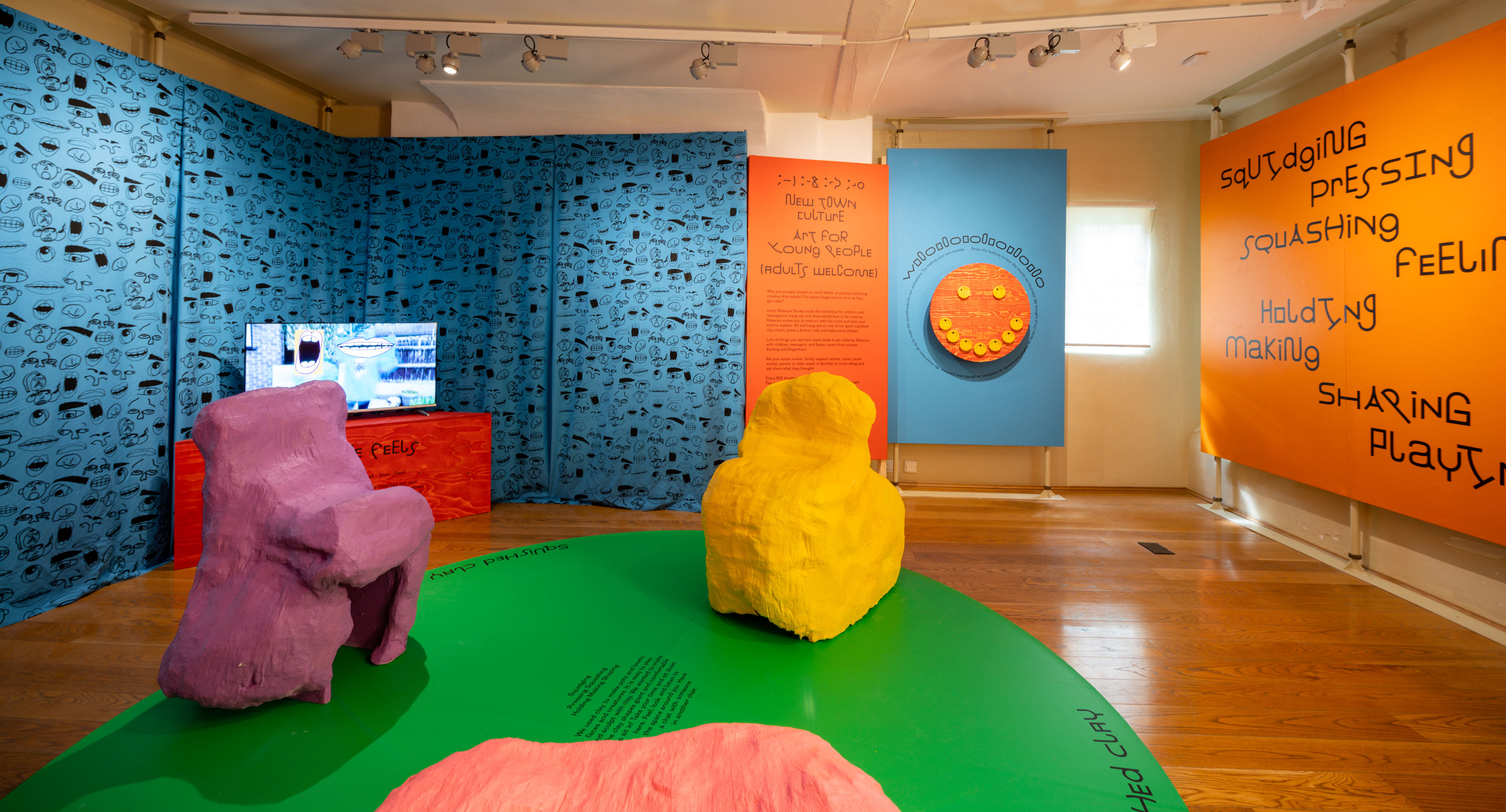 Exhibition space with bright blue wall hangings, orange painted walls, large screens showing art films and two very large purple and yellow chairs