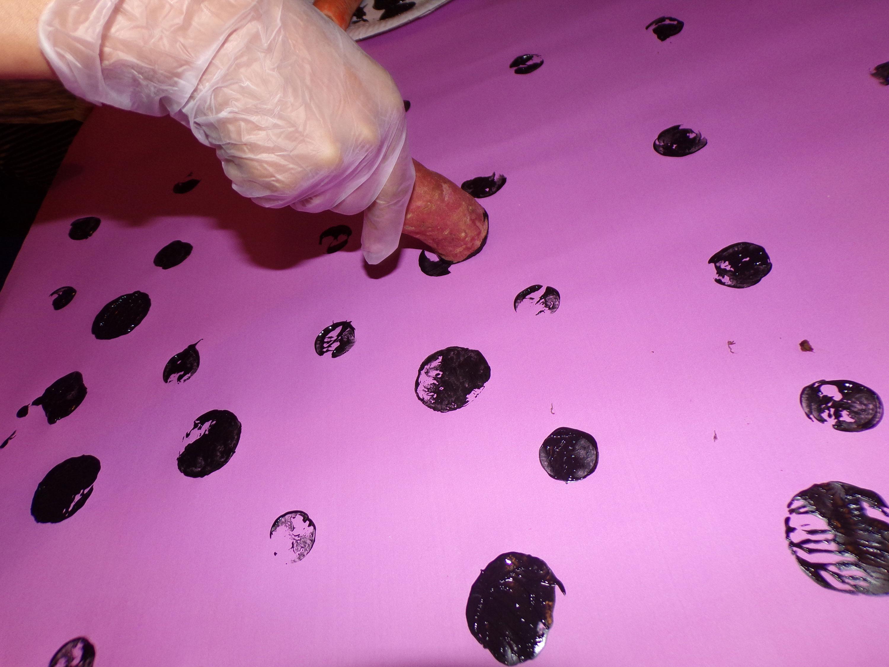 A close-up of a hand using a carrot to create a pattern of black dots on pink paper