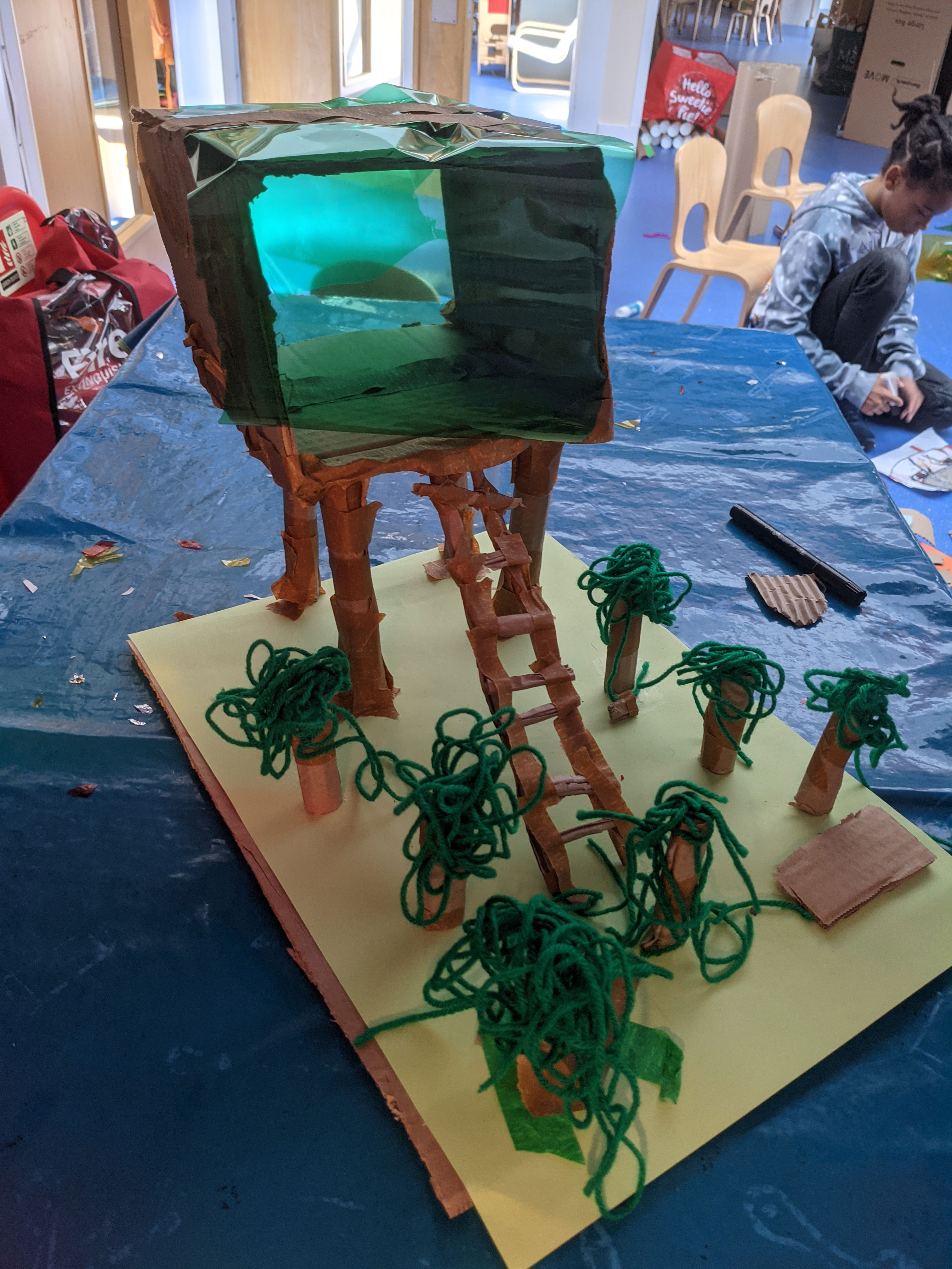 A small treehouse made with cardboard, tape, string and acetate placed on a sheet of A4 paper