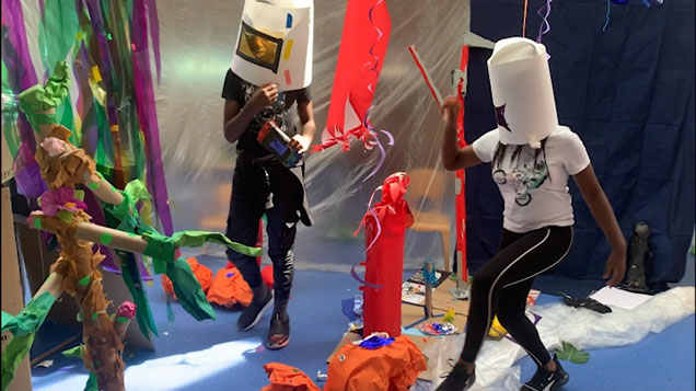 Moving image still of two participants wearing paper masks exploring the hanging and static materials