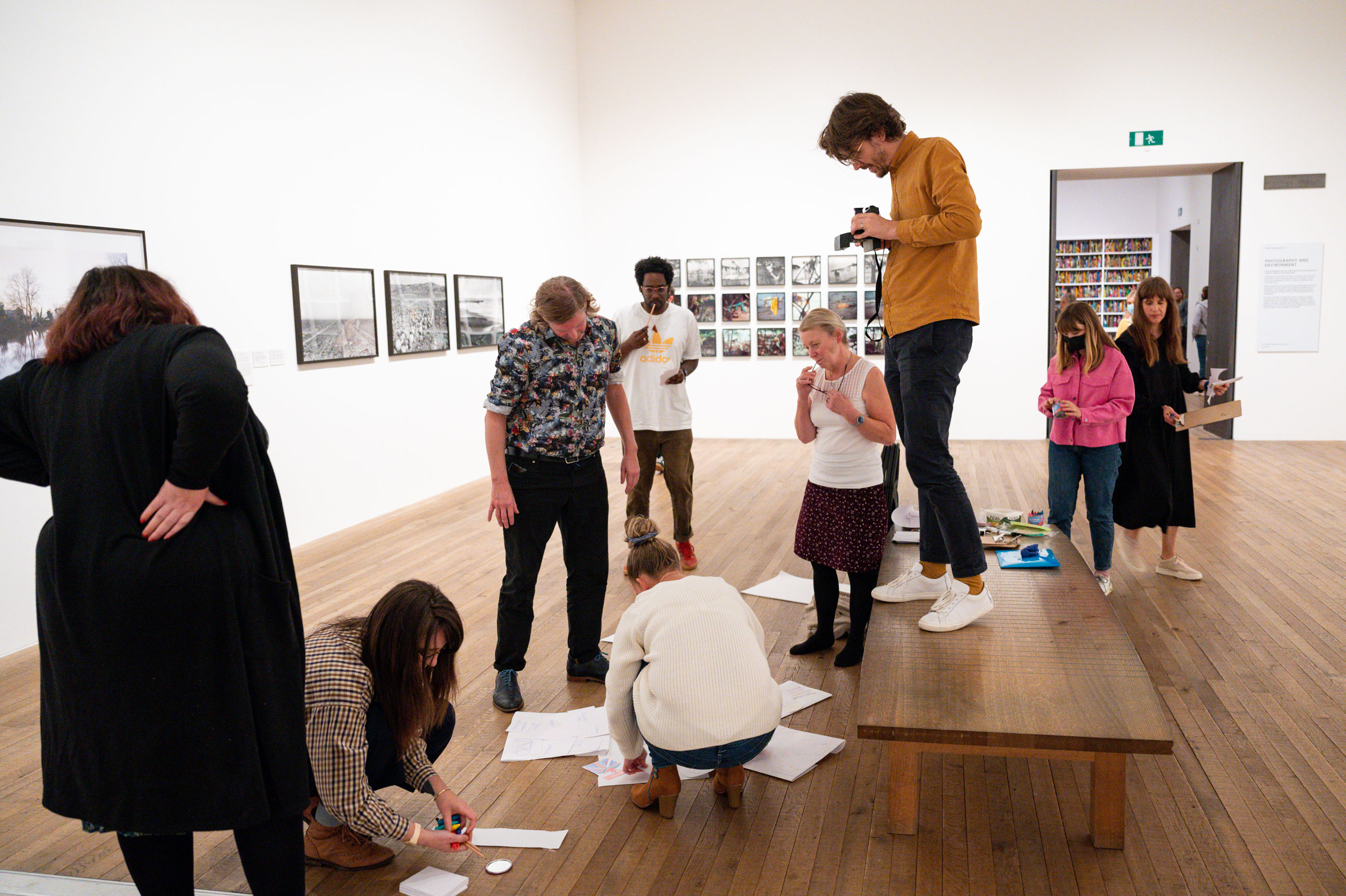 A group of people partaking in an activity in an exhibition space. Some people sitting on the floor, standing and standing on a stool