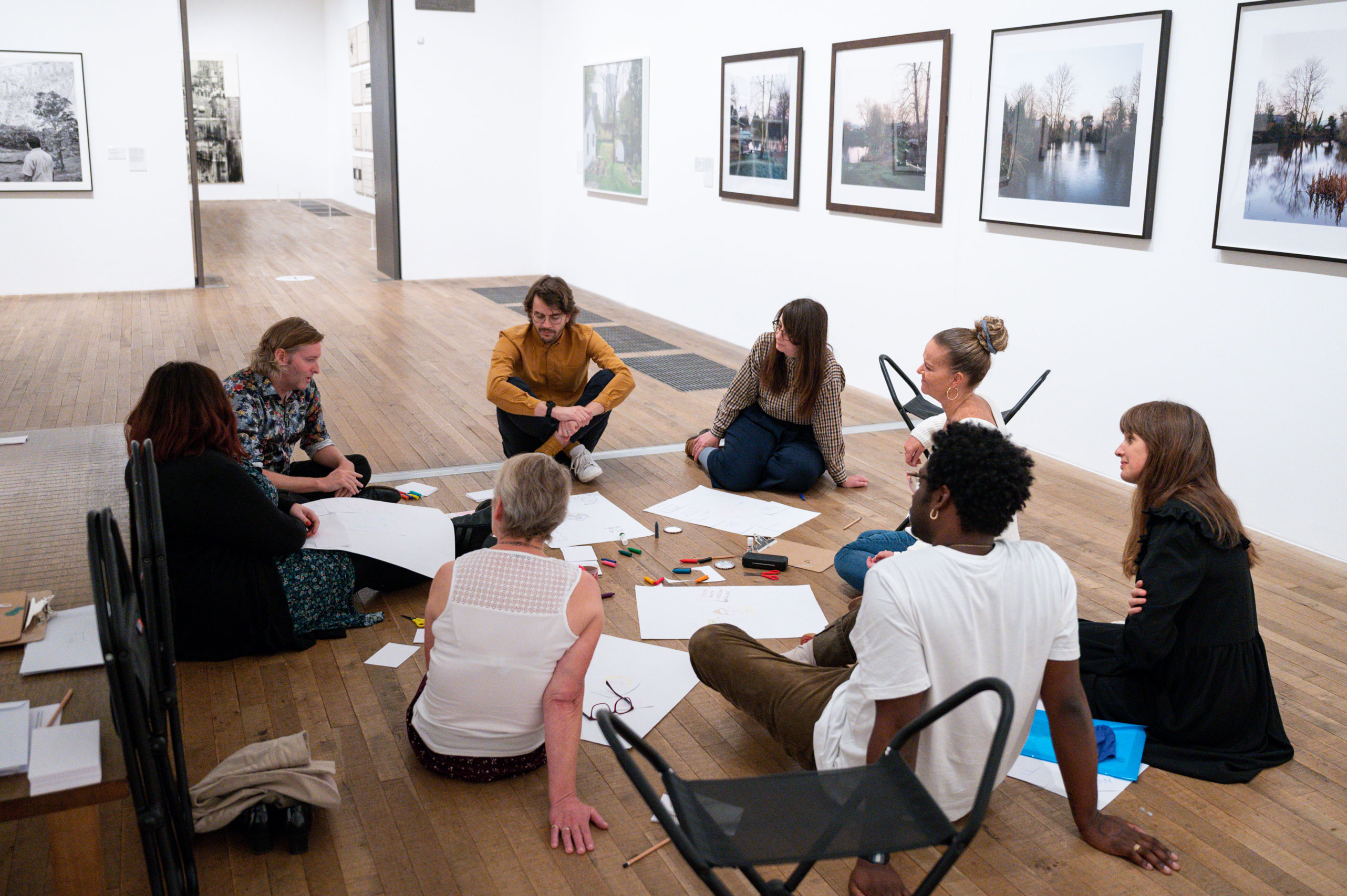 A group of people sitting on the floor of an art gallery surrounded with paper and pencils