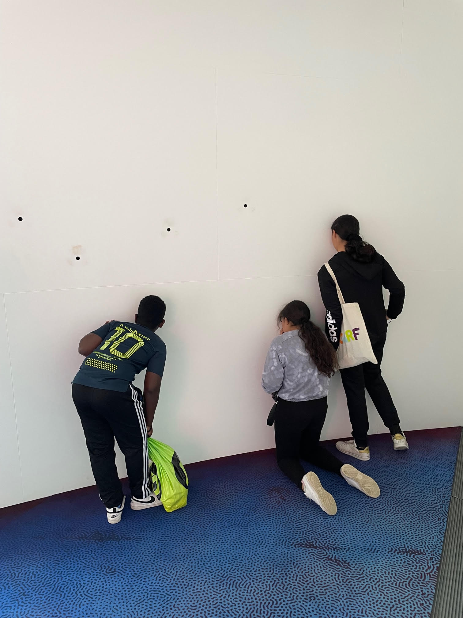 3 young people peering through small holes in a wall in an exhibition space