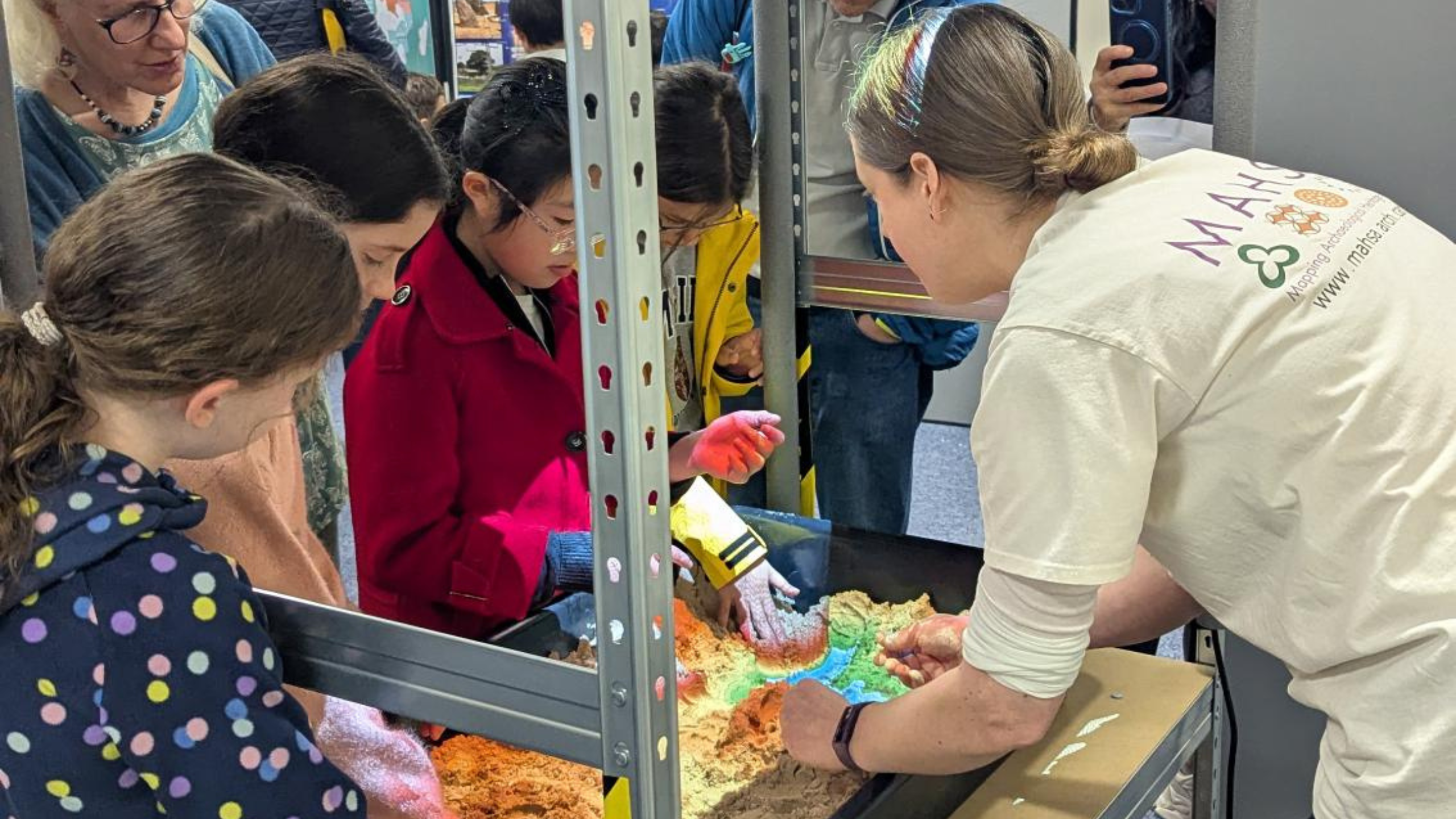 A photograph of a workshop leader demonstrating an AR sand box, with children watching around her and playing with the sand.